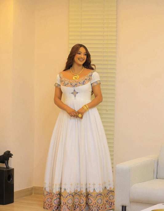 Woman in a white Habesha Dress with floral patterns standing in a room with a couch and decorative items.