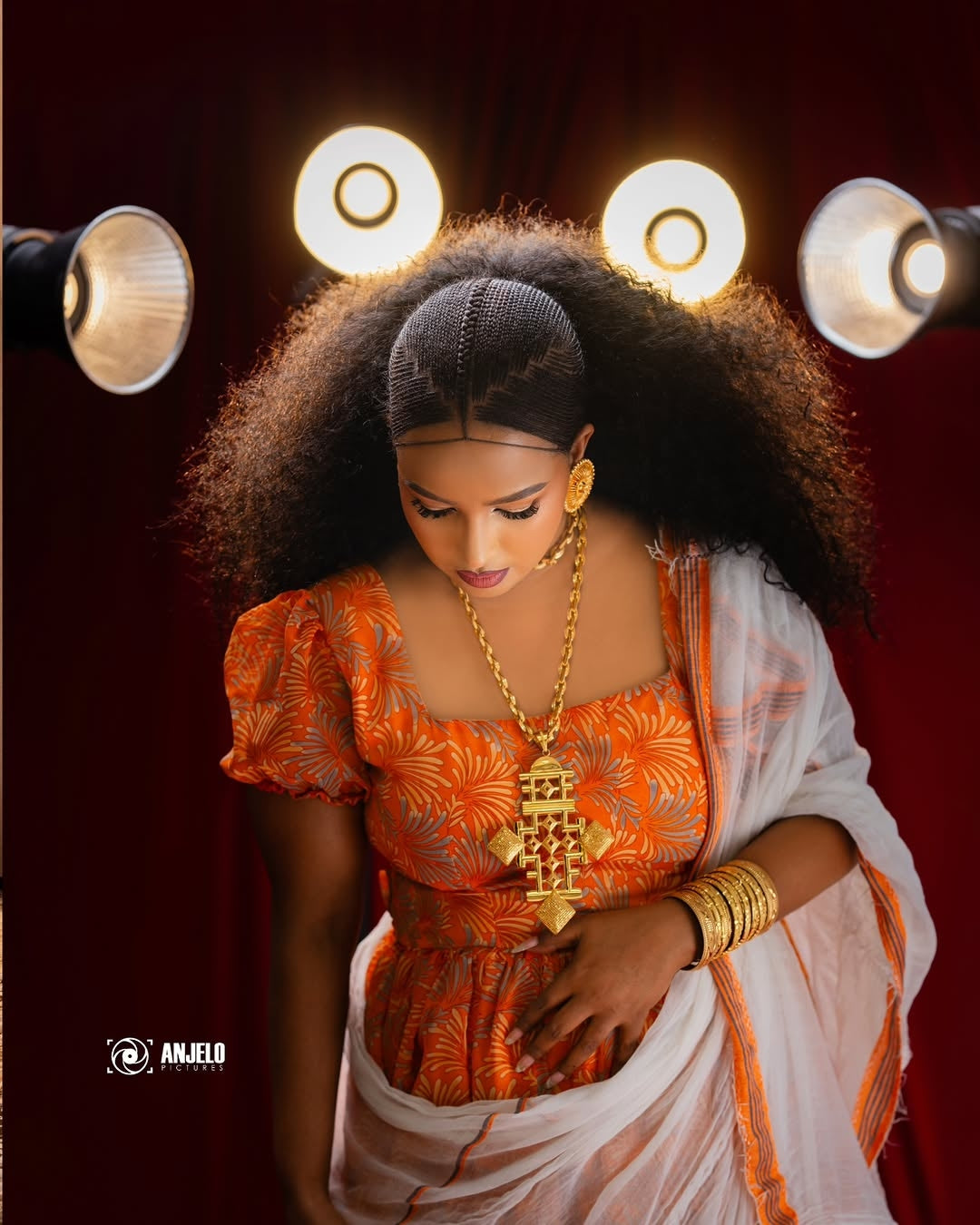 Woman in traditional Orange Habesha Chiffon with gold jewelry against a dark background
