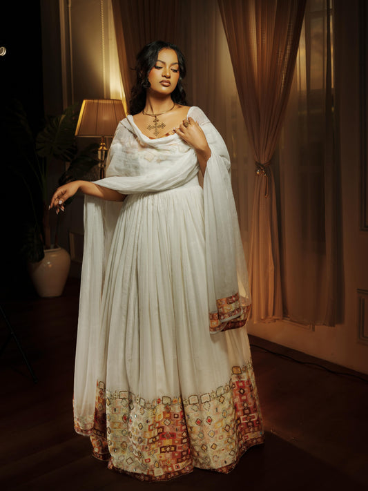 A woman models a traditional white Ethiopian national dresses with a matching dupatta and a heavily embroidered multicolor border at the hem, set against a warm, dimly lit background with curtains and a lamp.