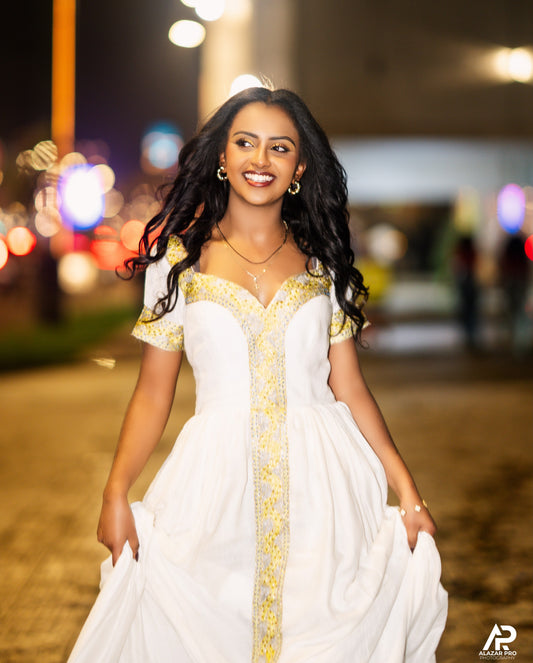 A woman wearing a white evening Ethiopian national dresses with gold embroidery and gold jewelry, smiling and posing outdoors at night with blurred city lights in the background.