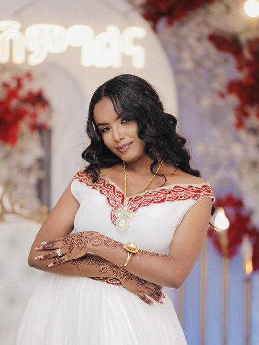 Woman in a white Ethiopian Traditional Dress with red embroidery standing in a decorated room.