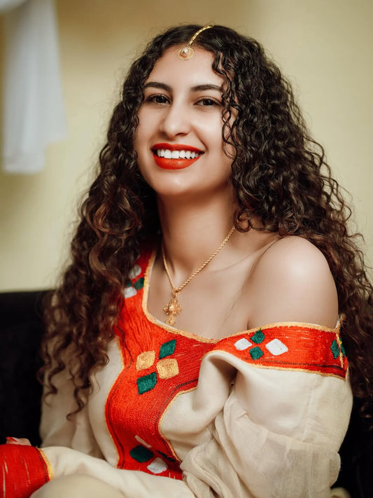 Woman wearing a Ethiopian traditional dress with a red blouse and beige dupatta, smiling.