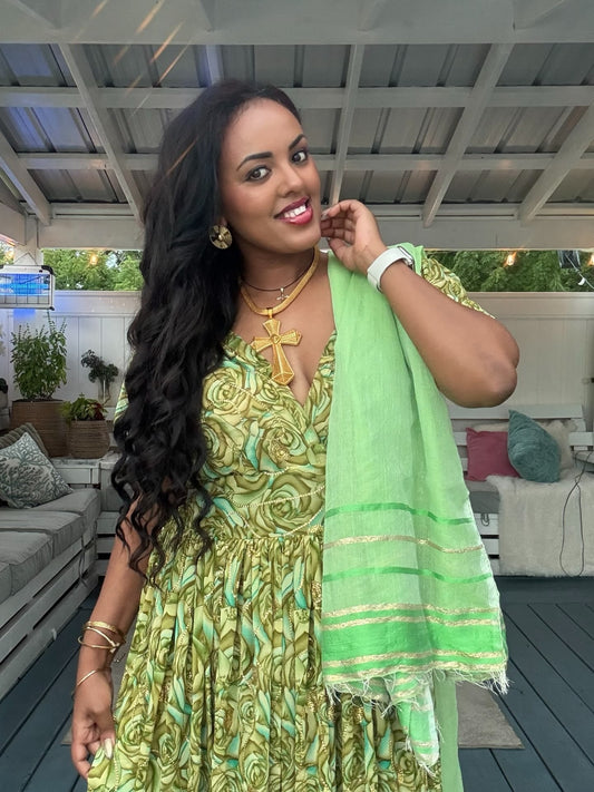 A woman wearing a green and gold patterned Habesha chiffon with a matching light green dupatta draped over her shoulder, standing on an outdoor patio.