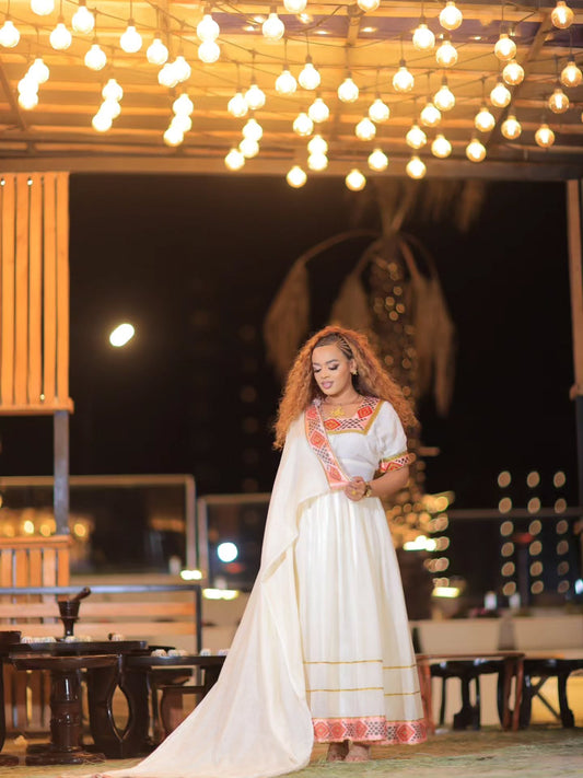 A woman wearing a long white Ethiopian Store Dress with orange and yellow embroidered details stands under a canopy of warm hanging string lights at night.