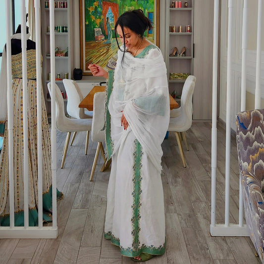 A woman wearing a white saree with green border Traditional Ethiopian Clothing stands in a home showroom or studio with light wood floors, white chairs, and shelving in the background.