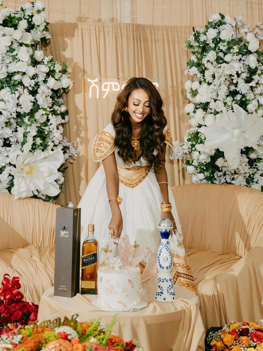 Woman in a white and gold Royal Gold Habesha Wedding Dress standing behind a table with a cake and bottles, decorated with flowers and lights.