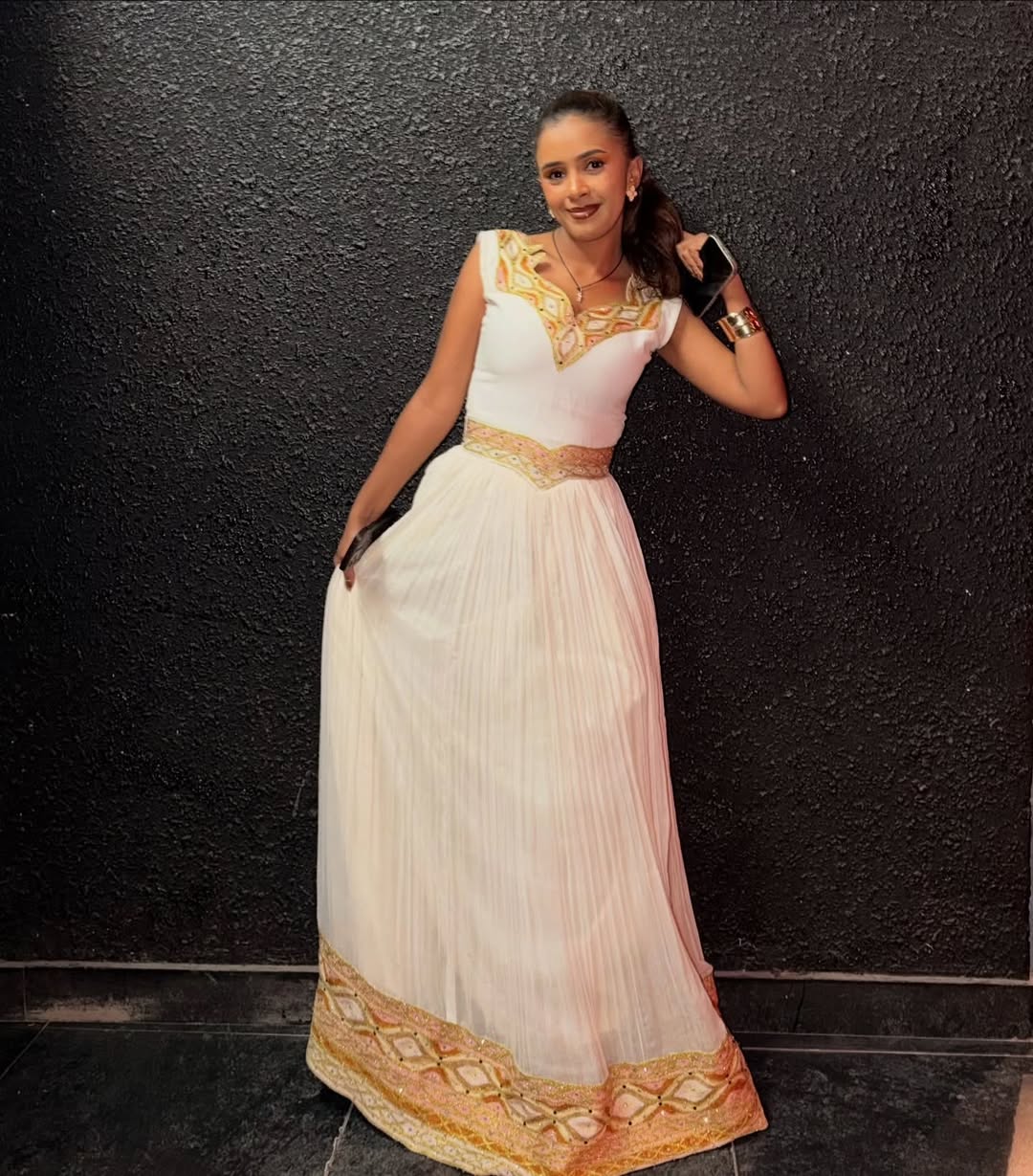 Woman in a white and gold traditional Ethiopian traditional dress against a black wall