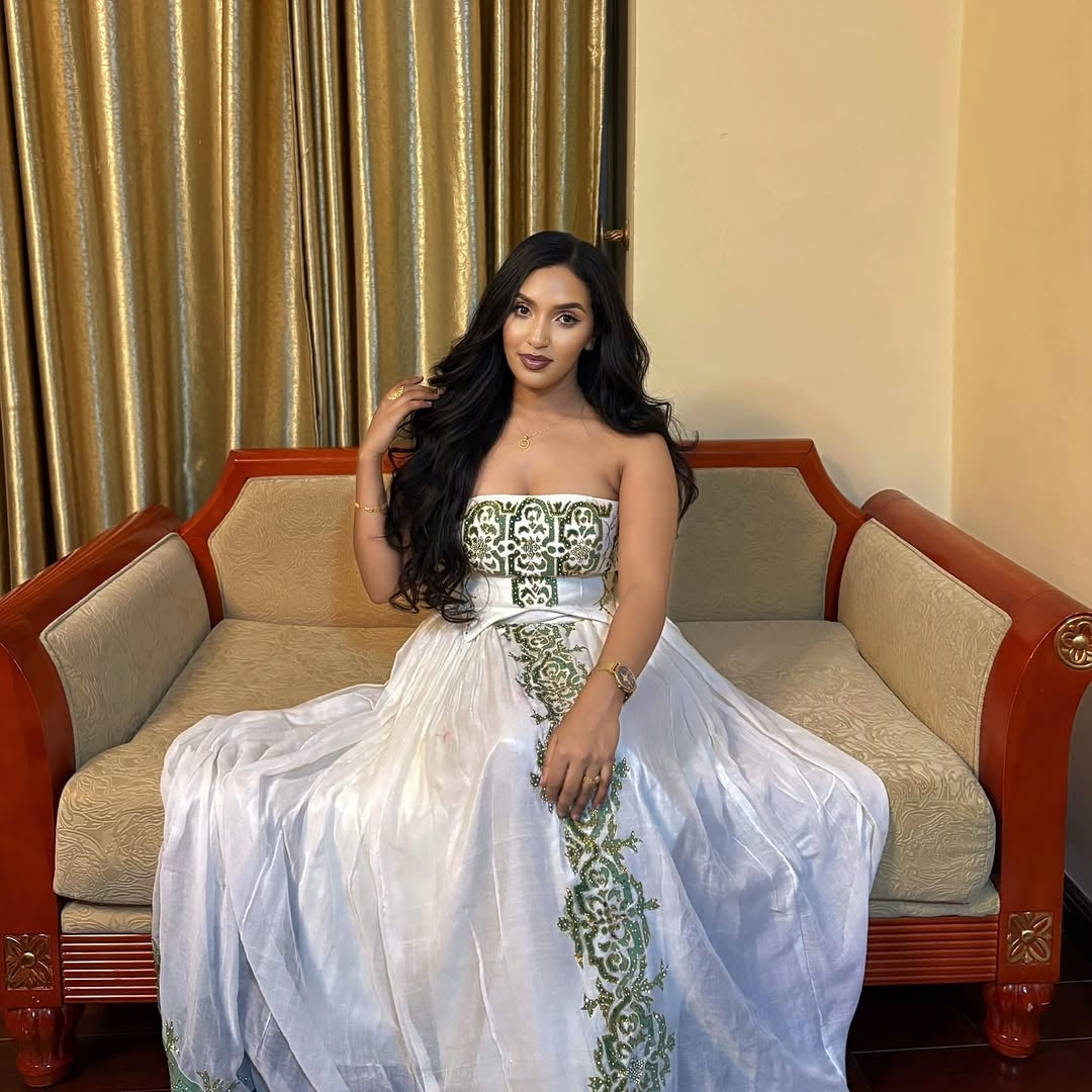 Woman in a white and green Ethiopian dress sitting on a red couch with gold curtains in the background
