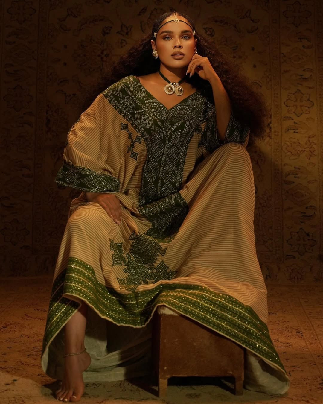 Woman in Gondar Fetel attire sitting on a wooden stool against a textured wall.