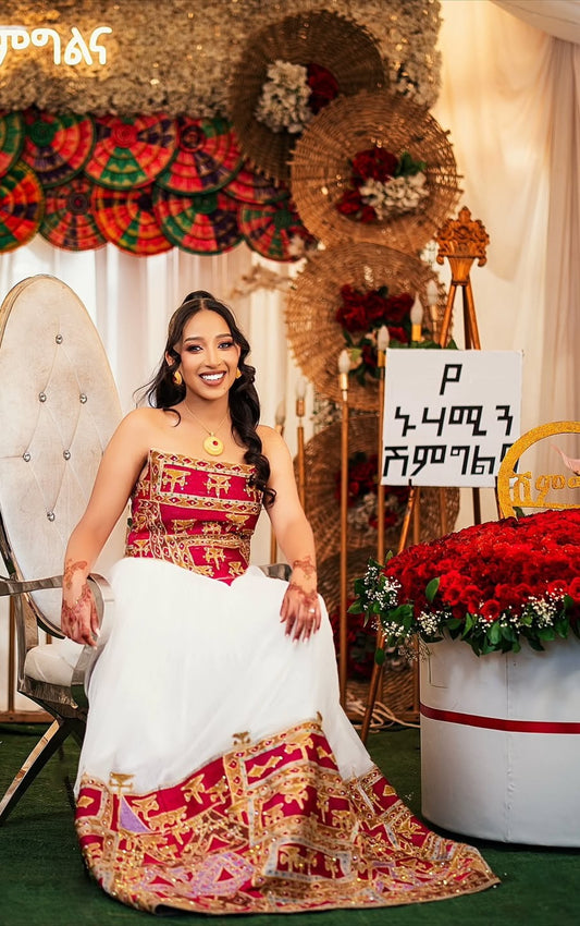 Woman in Habesha Wedding outfit sitting in a decorated room with floral arrangements and a chair.