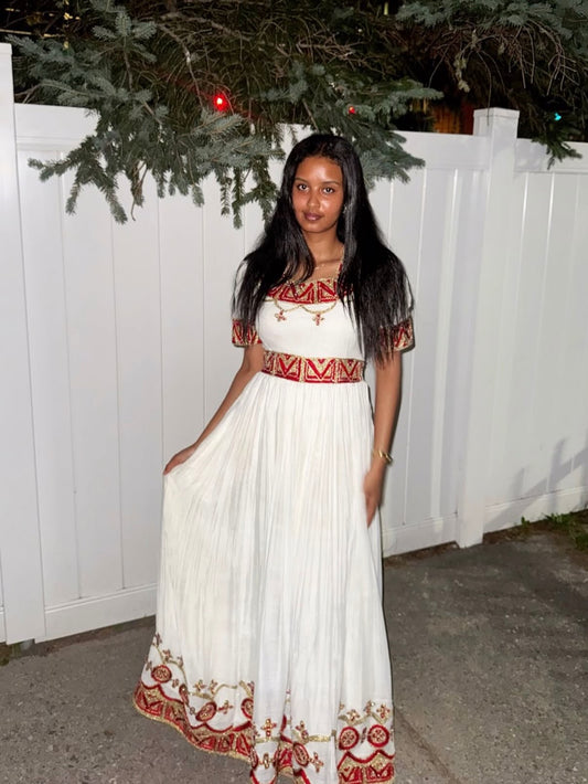 A woman wearing a long white maxi Habesha Zuria with red and gold geometric embroidery, standing in front of a white fence at night.