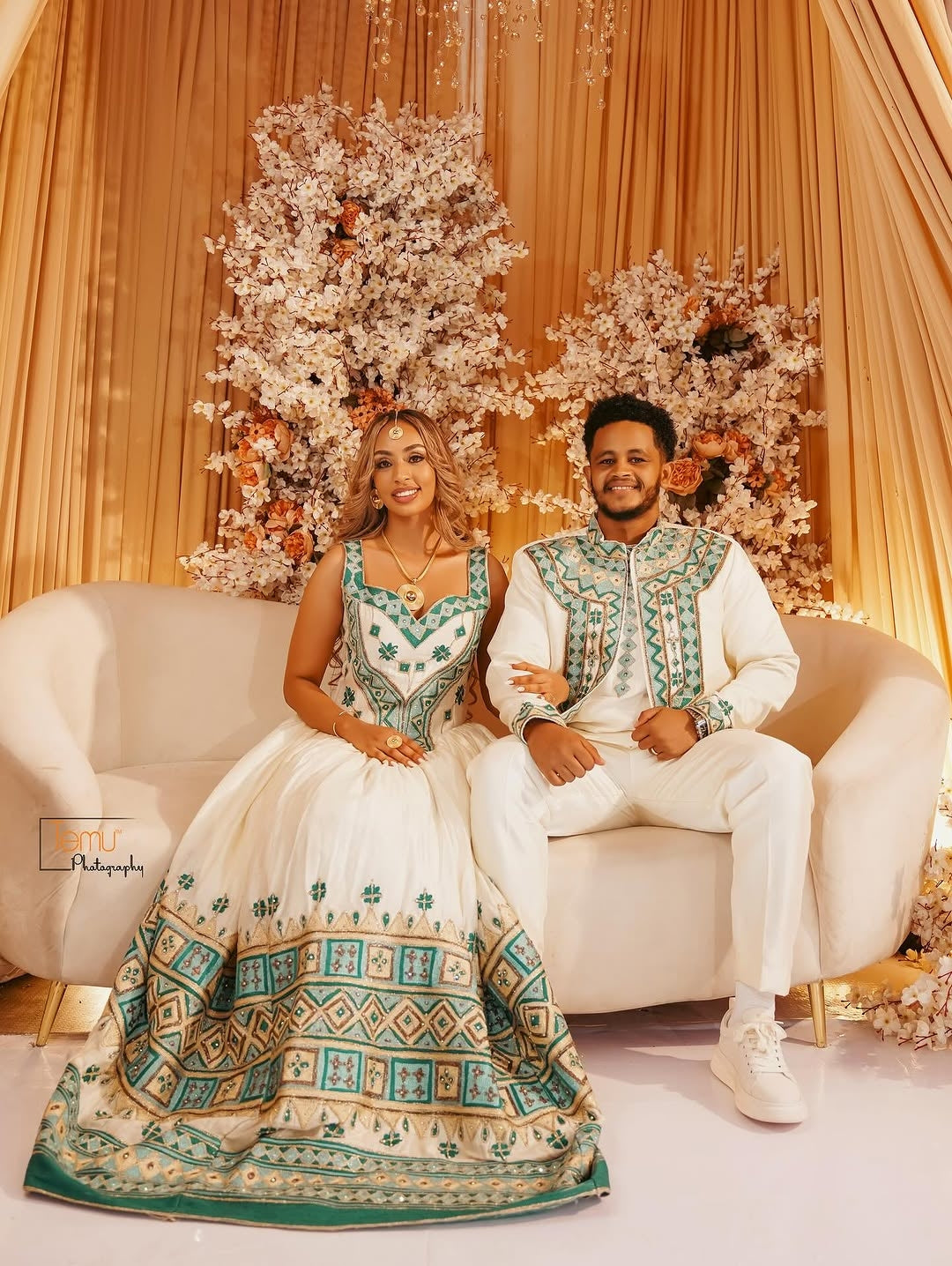 Couple in formal habesha wedding attire sitting on a white couch with floral decorations in the background.
