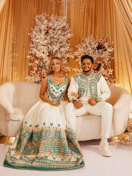 Couple in formal habesha wedding attire sitting on a white couch with floral decorations in the background.