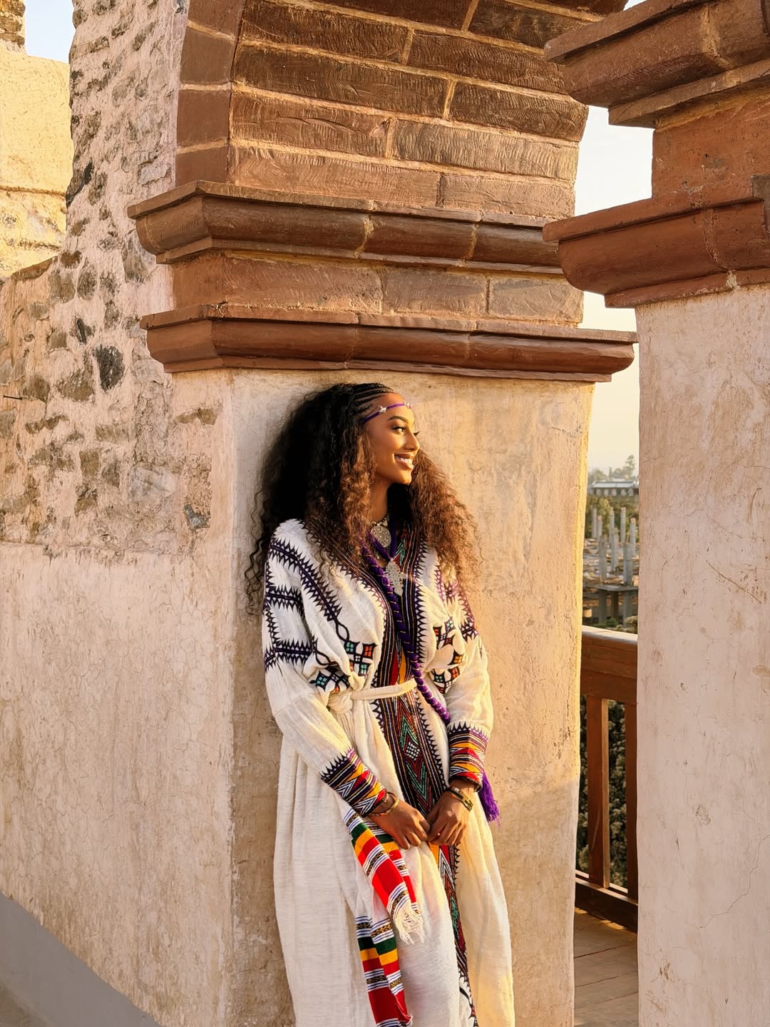 Woman in a long, patterned Ethiopian dress standing against a stone wall.