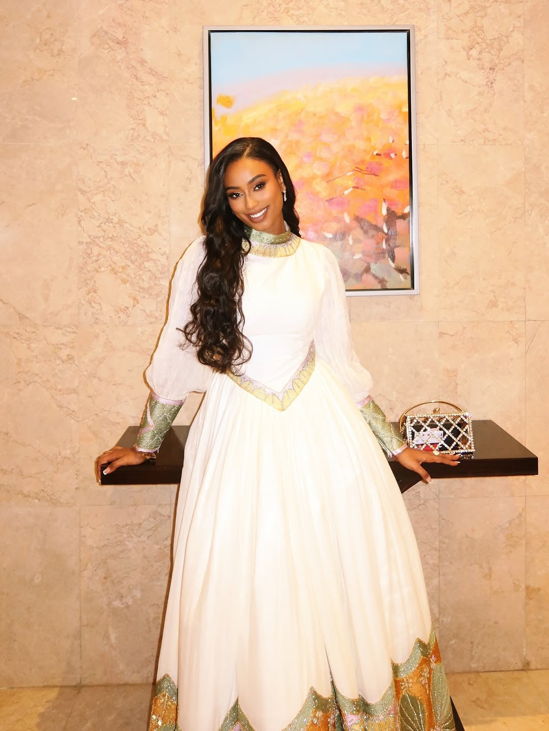 Woman in a white and green Habesha Dress standing indoors with a painting in the background.