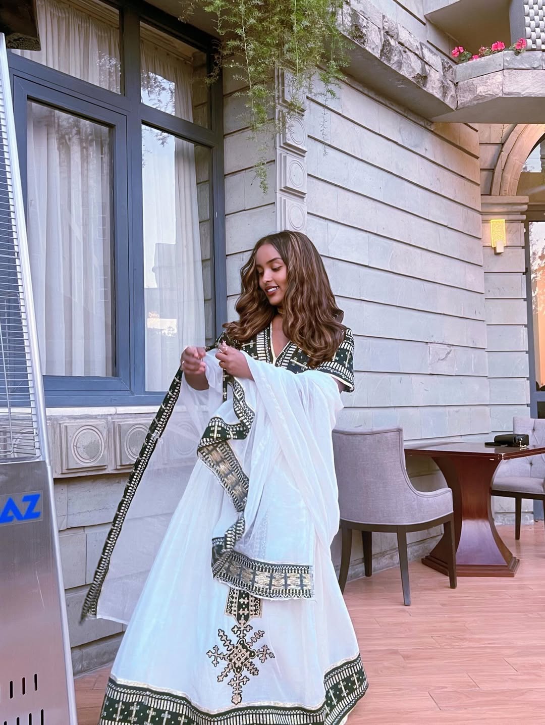 Woman in a white and green traditional Ethiopian traditional dress standing in an outdoor setting with a building and table in the background.