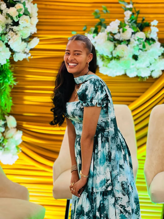 Woman in a floral Green Habesha Chiffon standing in front of a decorative floral backdrop with yellow accents.
