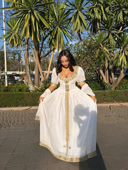 Woman in a white Habesha Zuria with gold accents standing outdoors with palm trees in the background