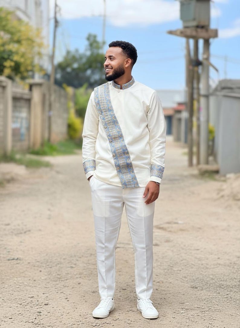 Man in a white Habesha Men's Outfit with a patterned sash standing on a street.
