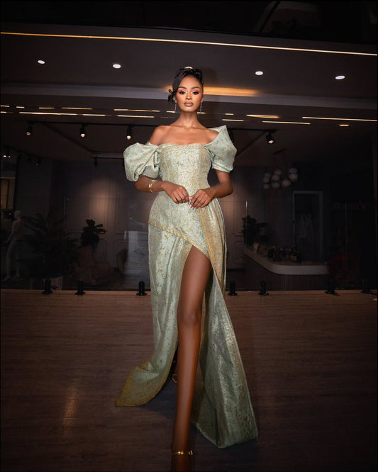 A woman models a floor-length, off-the-shoulder Habesha Wedding Dress with a high thigh slit in a dimly lit indoor venue. The dress has a shimmering pale sage green and gold textured fabric with dramatic puffed sleeves.