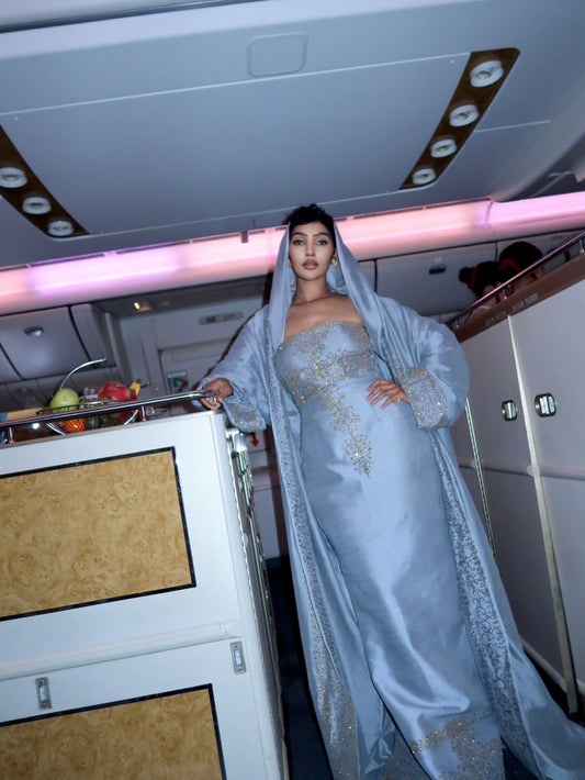 A woman wearing a light blue traditional Habesha Wedding Dress with gold embroidery and a matching head covering, standing inside an airplane cabin next to a service cart.