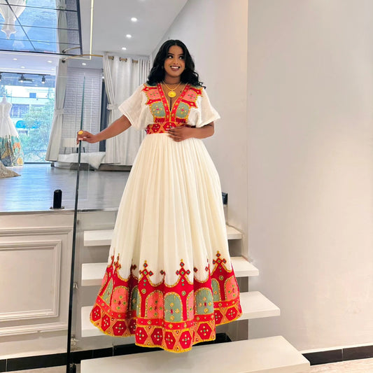 A woman wearing a traditional long white habesha Wedding dress with vibrant red, green, and yellow embroidery, standing on white stairs inside a modern boutique.