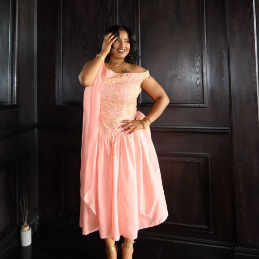 A woman models a peach-pink off-the-shoulder pink Habesha Wedding Dress with gold embroidery and beadwork, standing in front of a dark wood paneled wall.
