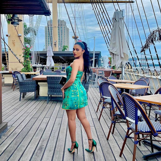 A woman stands on a wooden deck at a nautical restaurant, wearing a strapless turquoise green mini Habesha Wedding Dress with gold embroidery and teal high heels.