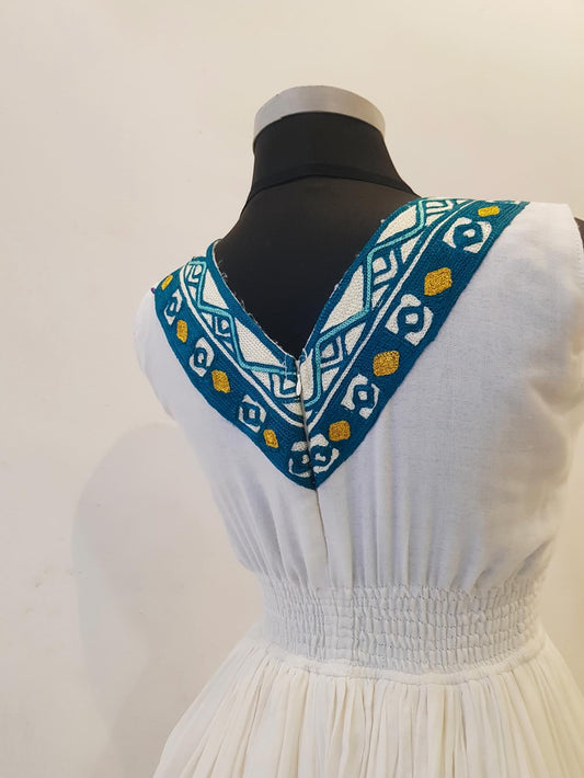 White sleeveless Ethiopian outfit displayed on a black mannequin torso, showing a V-neckline with a decorative embroidered border in blue, white, and yellow.