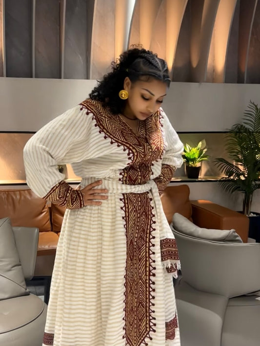 A woman wearing a long white Ethiopian traditional clothing with brown embroidered patterns and a matching belt, standing in a modern living room setting.