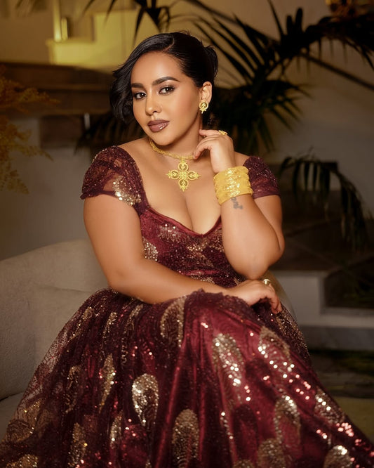 Woman in a sparkling red and gold habesha chiffion dress with jewelry, sitting indoors.