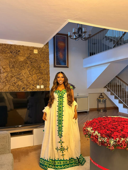 Woman in a green and white Green Habesha Libs standing in a modern living room with a large flower arrangement.