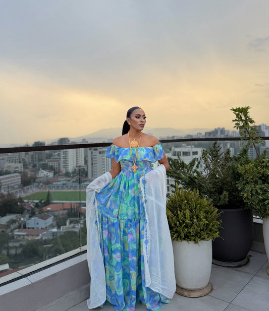 A woman stands on a rooftop balcony wearing a colorful off-the-shoulder maxi chiffion Habesha with a blue, green, and yellow abstract print, holding a sheer white wrap. The background shows a cityscape at dusk with buildings, mountains, and potted plants.