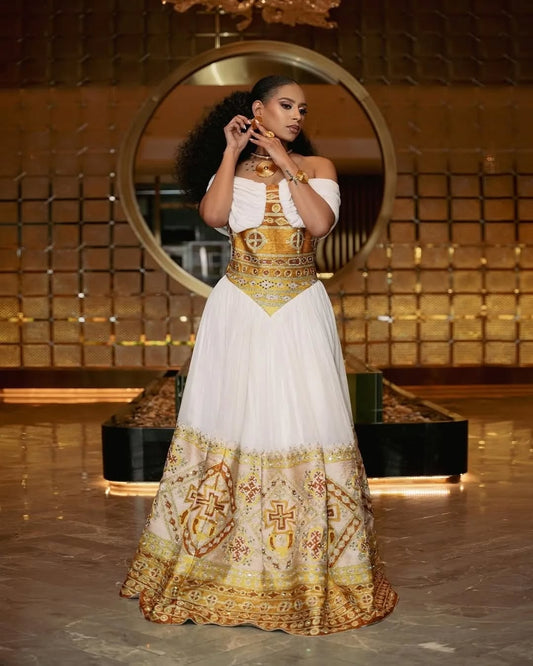 Woman in a white and gold traditional Gold Habesha Kemis standing in front of a decorative mirror.