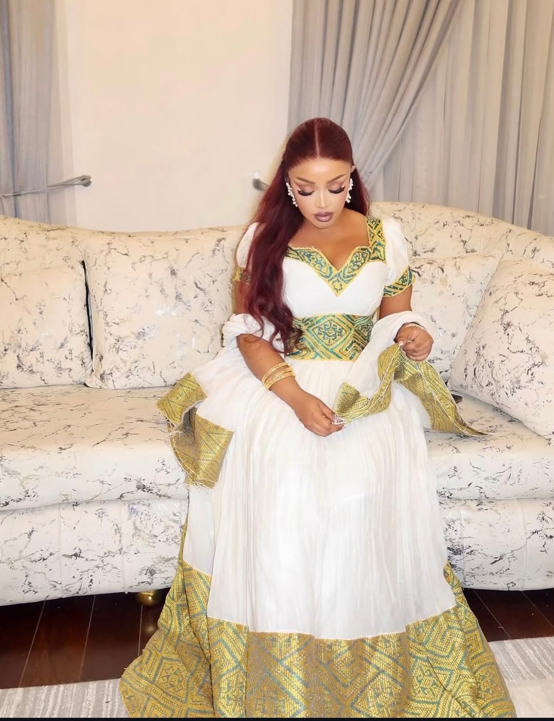Woman in a white and green Ethiopian traditional dress sitting on a patterned couch.