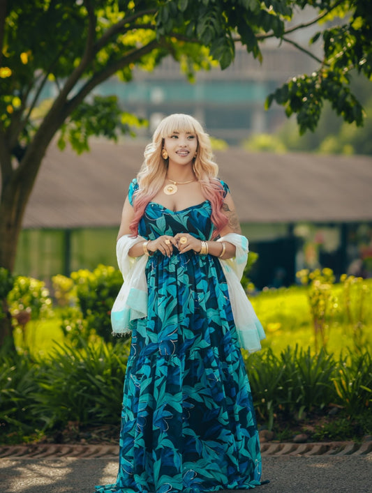 A woman stands outdoors in a garden wearing a long blue and teal leaf-patterned Habesha chiffion with white sheer sleeves and a white headpiece.