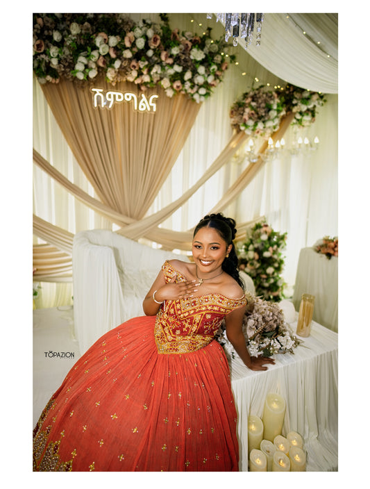 A woman wearing a voluminous off-the-shoulder red and gold traditional ball Habesha Wedding Dress poses in an elegantly decorated indoor event space with draped cream fabric, floral arrangements, and candles.