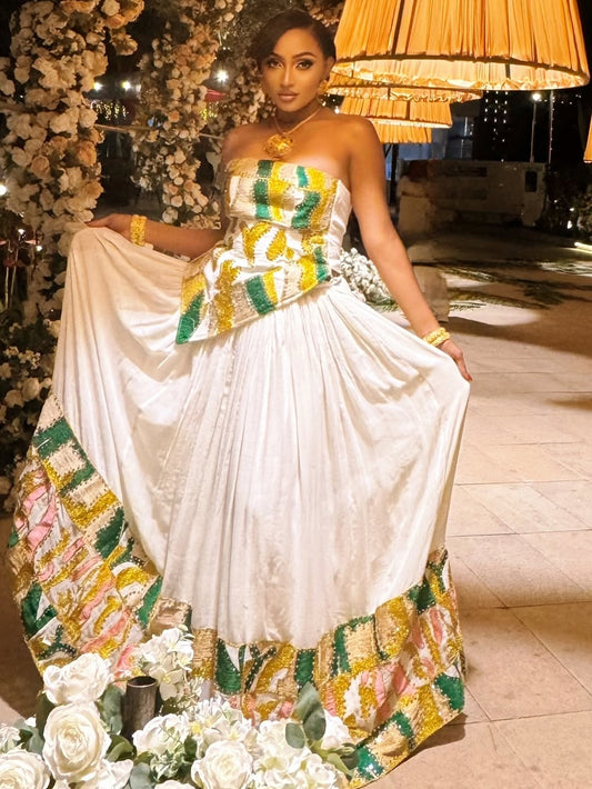 Woman in a white and colorful Habesha Dress with floral decorations, standing in an elegant indoor setting.