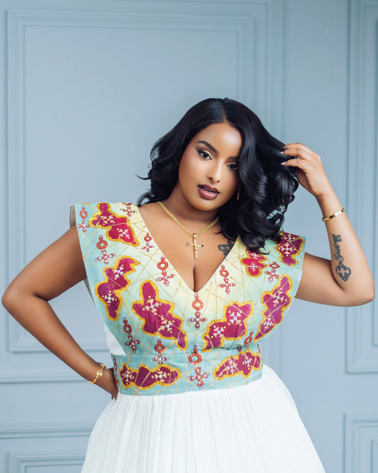 A woman modeling a sleeveless V-neck habesha Wedding dress with a light blue and white floral patterned bodice and a white skirt, posed against a light blue paneled wall.