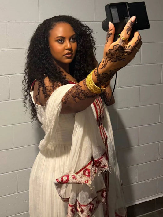 A woman taking a mirror selfie wearing a white sleeveless Habesha Kemis with red patterned trim and a matching white scarf, against a light gray brick wall.