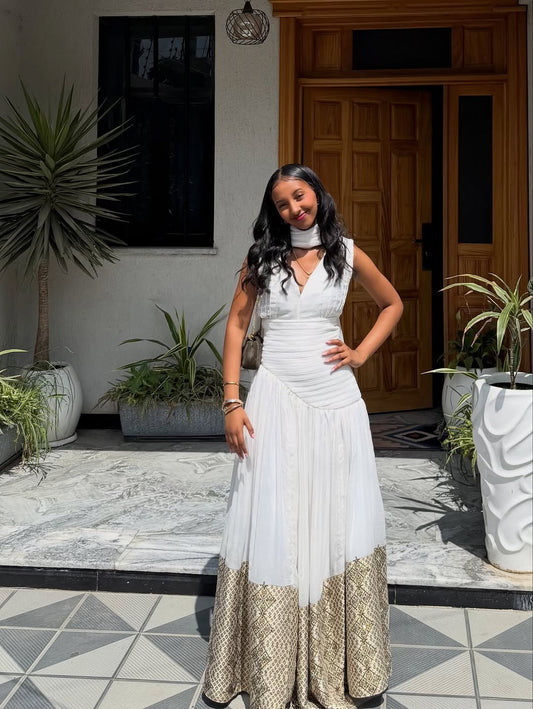Woman in a white and gold Habesha Dress standing in a decorative indoor setting with plants and a wooden door.