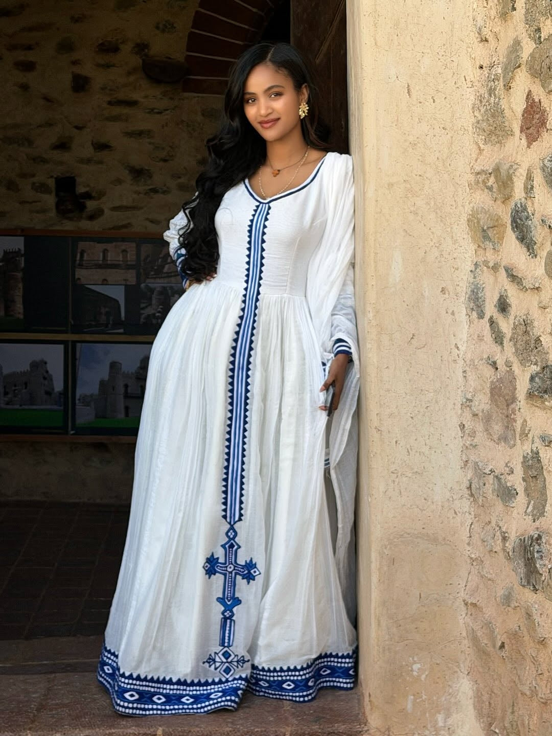 Woman in a white Ethiopian Dress with blue embroidery standing against a stone wall.