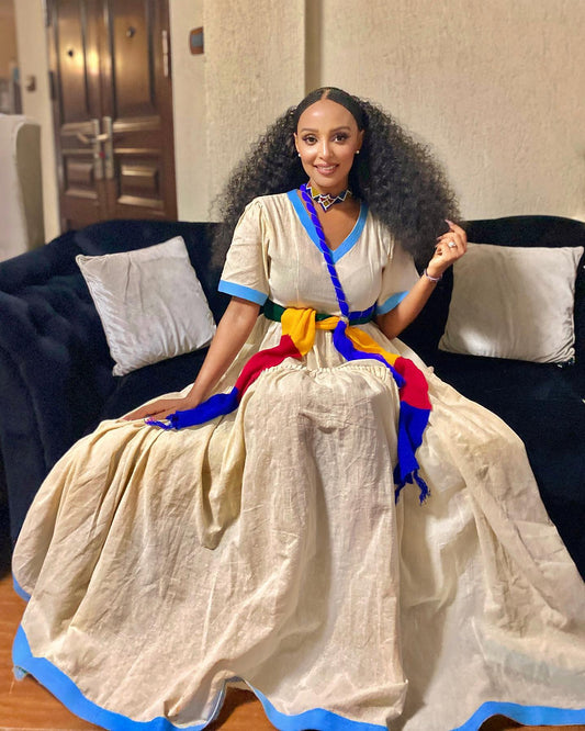 A woman with long, curly black hair sits on a black couch wearing a voluminous cream-colored Ethiopian National Dress with blue trim, a colorful striped sash, and a matching blue headpiece. The background shows a beige wall and a wooden door.