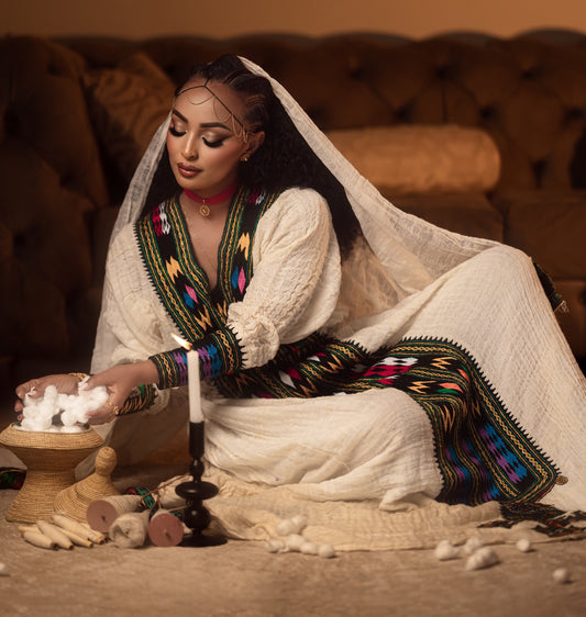 A woman in traditional West African attire, featuring a cream-colored embroidered Axum Fetil with vibrant multicolor geometric patterns, sits on the floor surrounded by cotton fibers, a lit candle, and wooden tools in front of a tufted brown sofa.