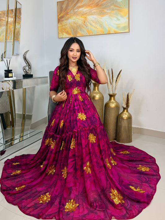 A woman models a long magenta tiered maxi Habesha Chiffon with gold floral patterns in an indoor setting with mirrors, gold vases, and a painting on the wall.