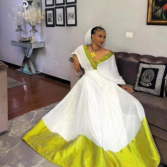 A woman wearing a white traditional Habesha Dress with a bright yellow-green border and matching headscarf, seated on a dark sofa in a living room.