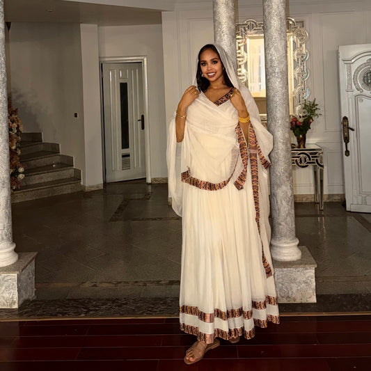 A woman stands in a grand indoor hallway wearing a traditional white Habesha Kemiswith brown patterned borders and a matching white dupatta.