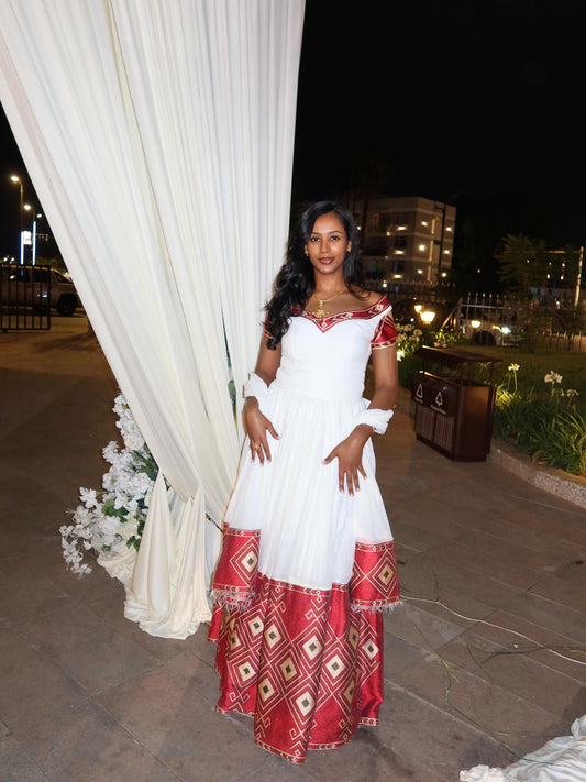 Woman in a white and red Habesha Kemis standing in front of a white curtain with flowers at night.