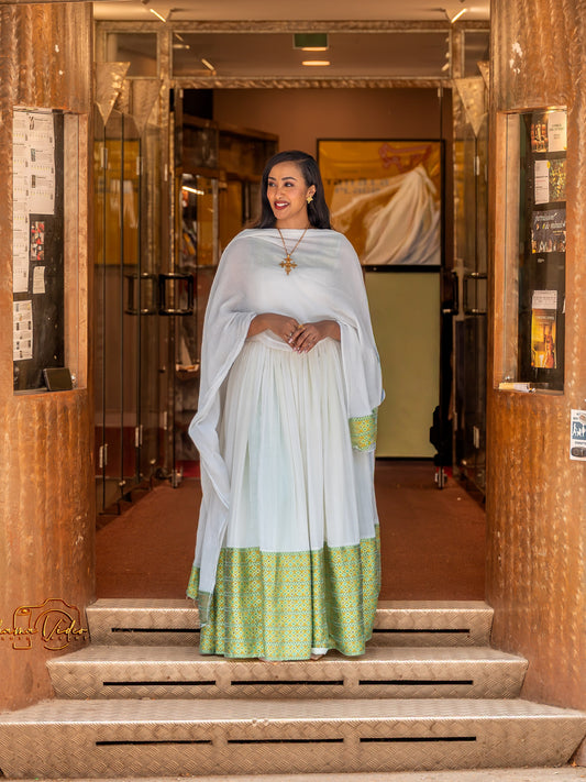 A woman stands on stone steps in front of a building entrance wearing a traditional white Ethiopian Dress with a light blue dupatta and a green patterned border, along with a gold cross necklace.