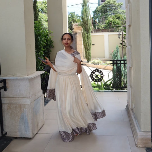 Woman in a white Habesha dress with brown patterns standing in an outdoor setting.